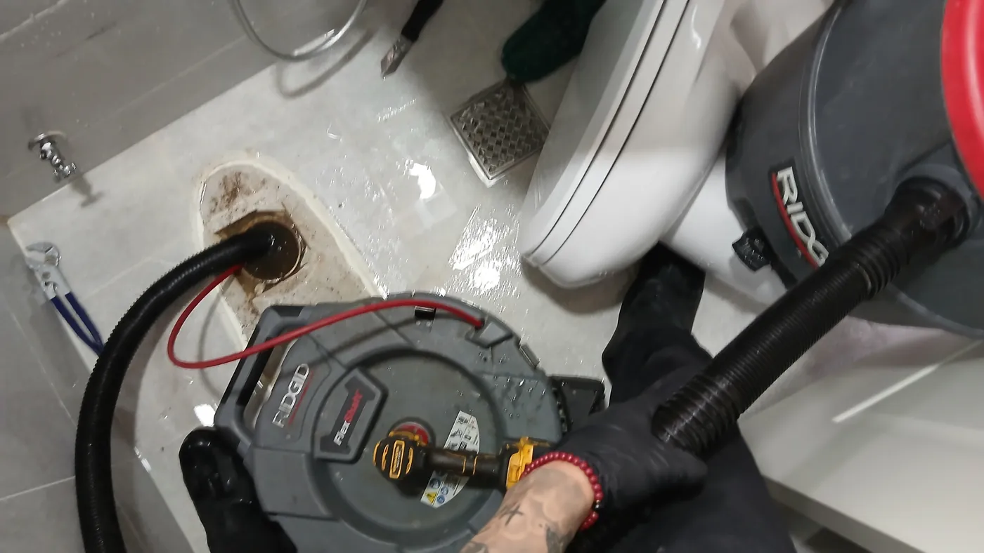 바닥 배관 입구로 들어가는 RIDGID 플렉스샤프트와 DeWalt 드릴, 옆에 떼어 둔 양변기