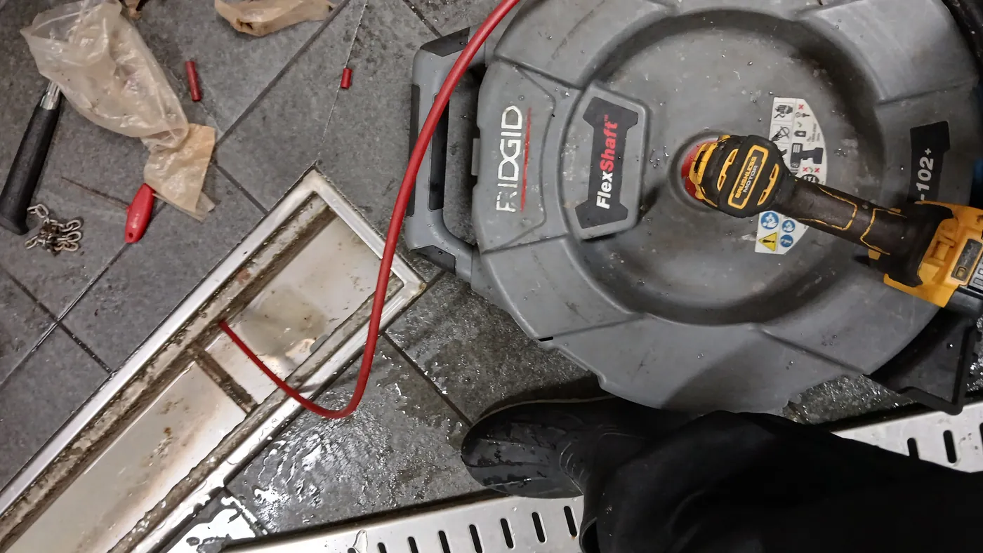 RIDGID FlexShaft K9-102 드럼과 충전 드릴, 붉은색 케이블을 주방 스테인리스 트렌치 배수로에 진입시킨 모습과 바닥의 체인 커터 헤드